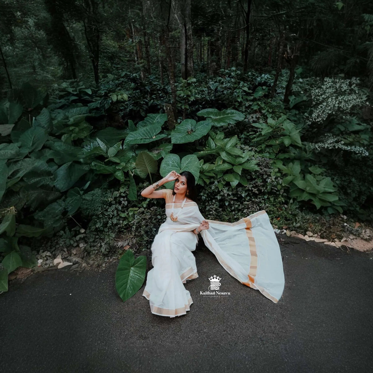 Woman in traditional white and gold Onam saree sitting by lush green foliage outdoors