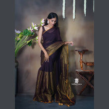 Woman in a dark purple saree with gold border standing next to a vase of flowers against a neutral background.